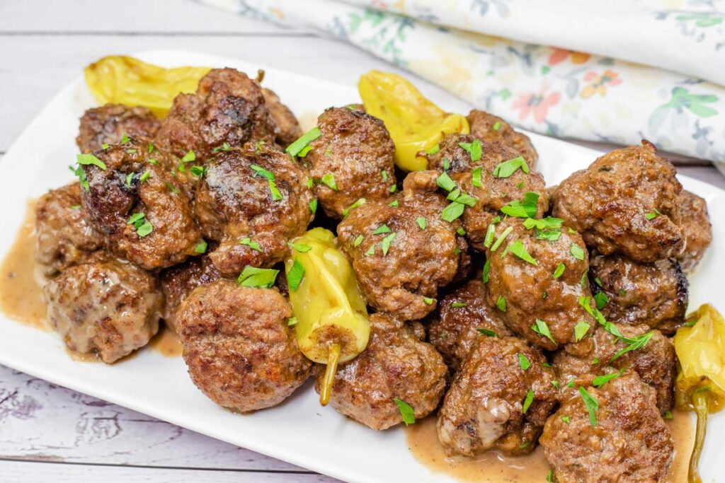 A plate of brown meatballs garnished with green herbs, accompanied by yellow peppers on a white rectangular dish.
