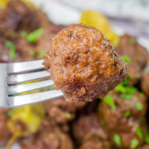 Close-up of a Slow Cooker Mississippi Meatballs on a fork with a plate of more meatballs in the background. Garnished with chopped herbs.