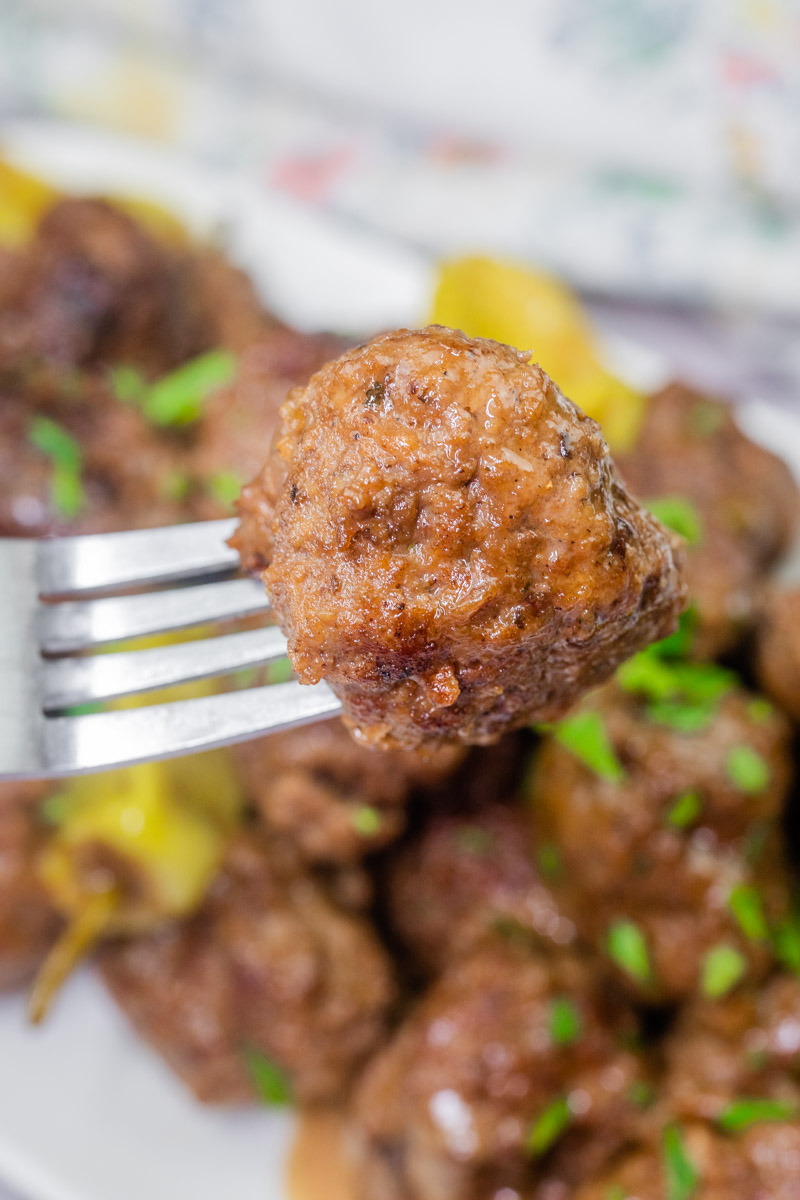 Close-up of a Slow Cooker Mississippi Meatballs on a fork with a plate of more meatballs in the background. Garnished with chopped herbs.