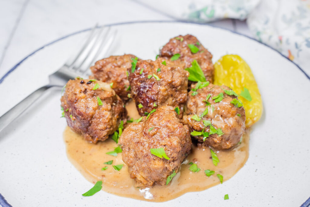 Plate of Slow Cooker Mississippi Meatballs with gravy, garnished with parsley, next to a green pepper. Fork and knife on the side.