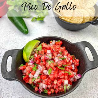 Bowl of pico de gallo with a lime wedge, placed on a table. Another bowl with tortilla chips and a jalapeño pepper are in the background.