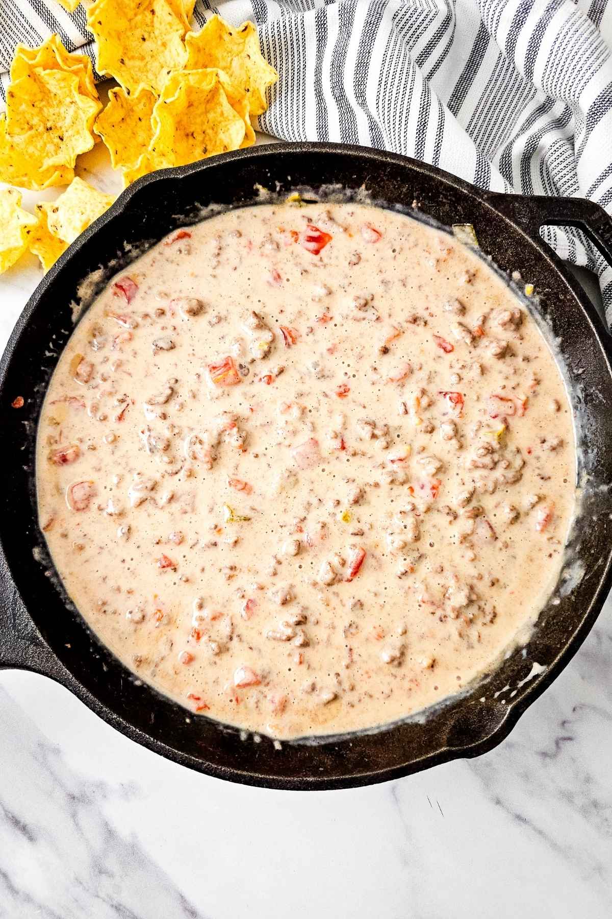 A skillet filled with creamy Queso Blanco Rotel Dip with visible diced tomatoes, surrounded by tortilla chips, on a marble surface with a striped cloth nearby.