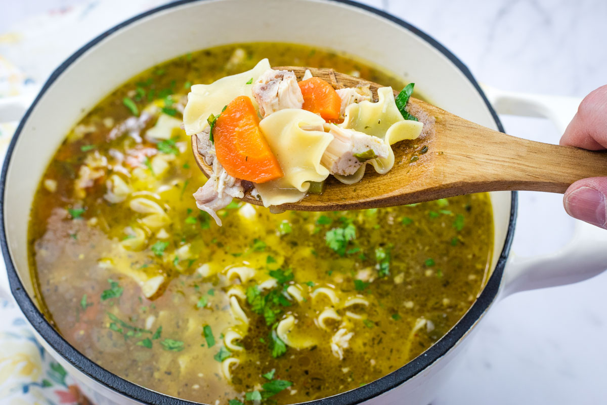 A wooden spoon holds Rotisserie chicken noodle soup above a pot filled with more soup.