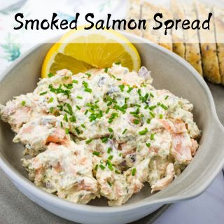 A bowl of smoked salmon spread topped with chopped chives, garnished with a lemon slice, next to sliced bread.