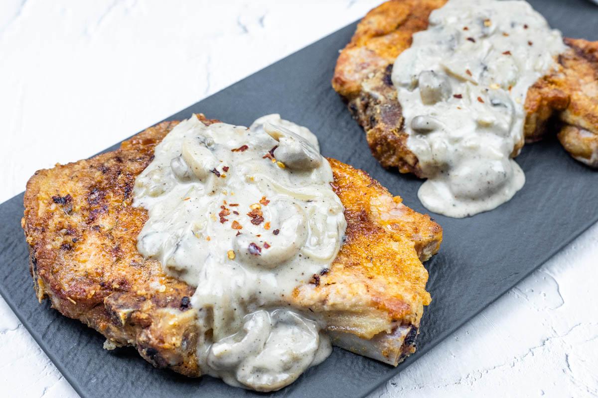 Two Saucy Smothered Pork Chops topped with creamy mushroom sauce on a black rectangular plate.