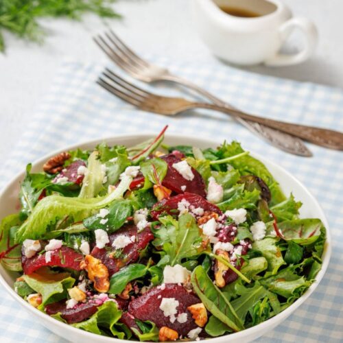 A salad with mixed greens, sliced beets, walnuts, and crumbled feta in a bowl, placed on a blue checkered cloth. Two forks and a small pitcher of dressing are in the background.