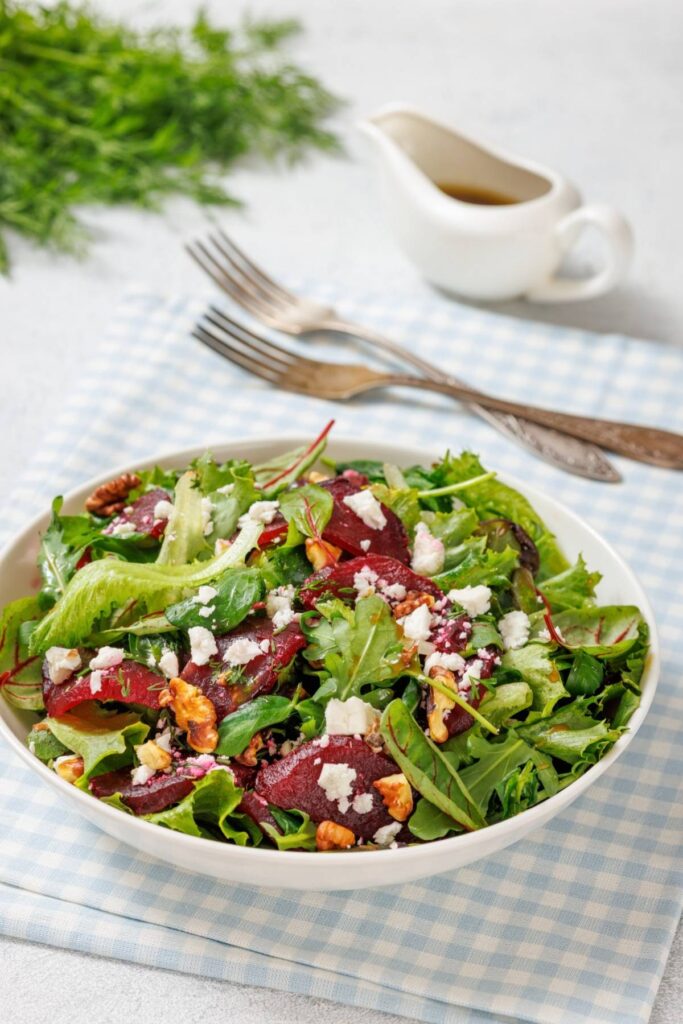 A salad with mixed greens, sliced beets, walnuts, and crumbled feta in a bowl, placed on a blue checkered cloth. Two forks and a small pitcher of dressing are in the background.