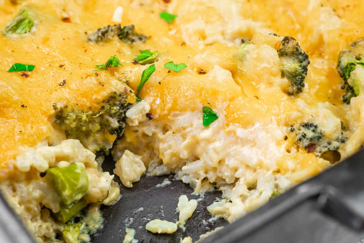Close-up of a cheesy broccoli and rice casserole in a baking dish, with a serving removed, showing melted cheese, rice, broccoli, and garnished parsley.
