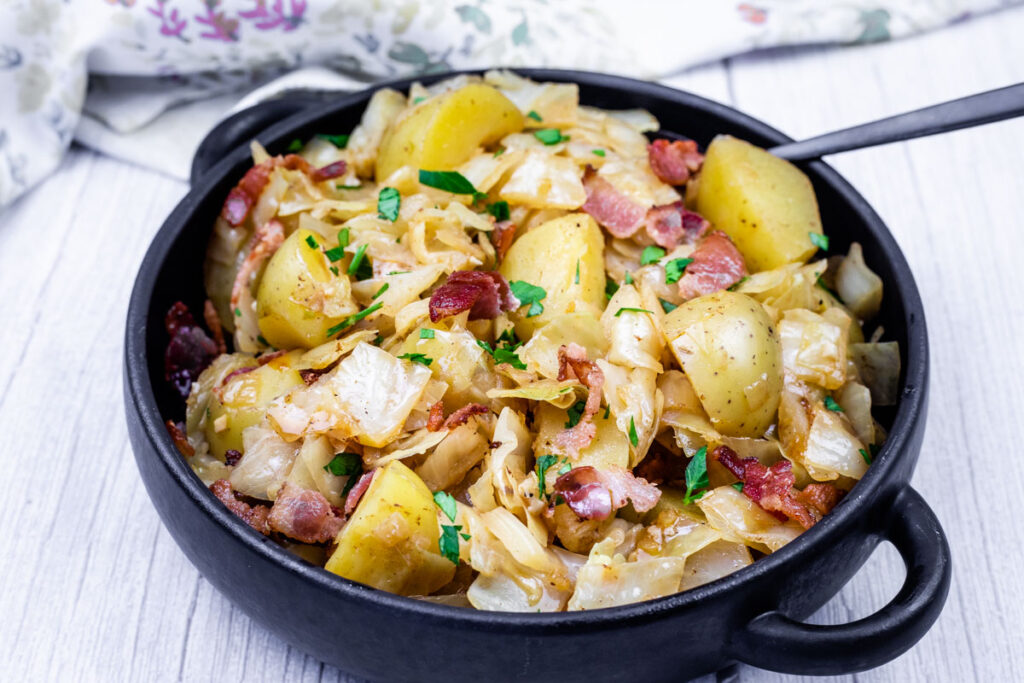 A black bowl filled with Cabbage & Potatoes with Bacon with a spoon on the side.