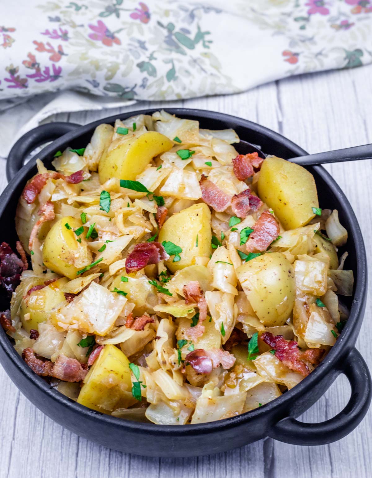 A black bowl of Cabbage & Potatoes with Bacon, garnished with chopped parsley. A floral-patterned cloth is in the background.