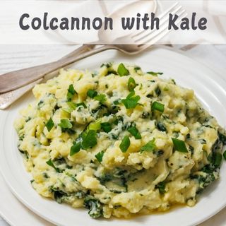 Plate of colcannon with kale, garnished with sliced green onions, accompanied by a fork and spoon on a napkin.
