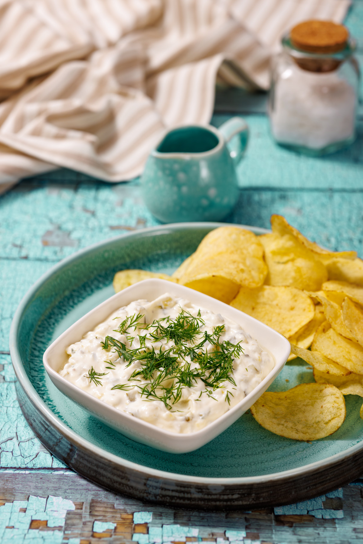 A bowl of Dill Pickle Dip topped with fresh dill, served with potato chips on a blue plate. A small pitcher and a jar of salt are in the background.