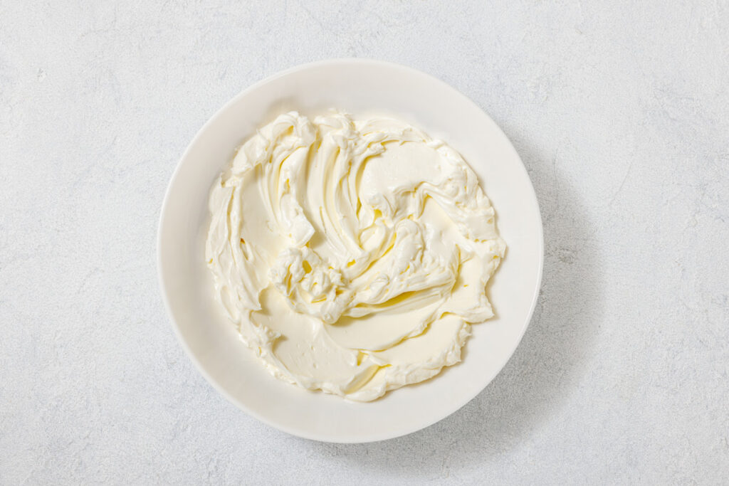 A white bowl filled with smooth, swirled cream cheese on a light, textured surface.