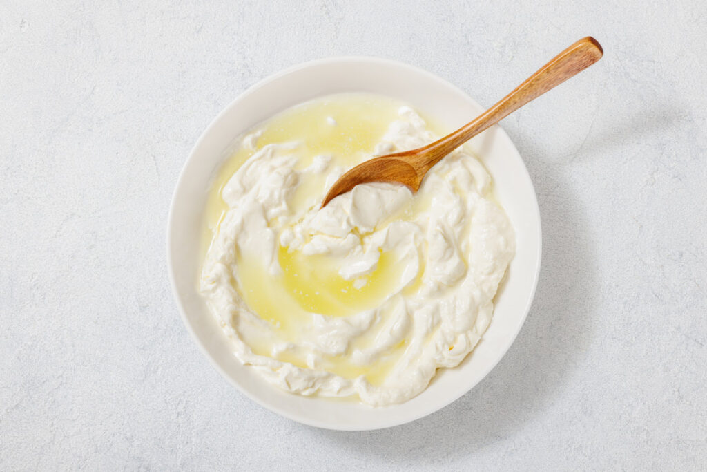 A bowl of yogurt with a drizzle of olive oil, and a wooden spoon resting on top, against a plain white background.