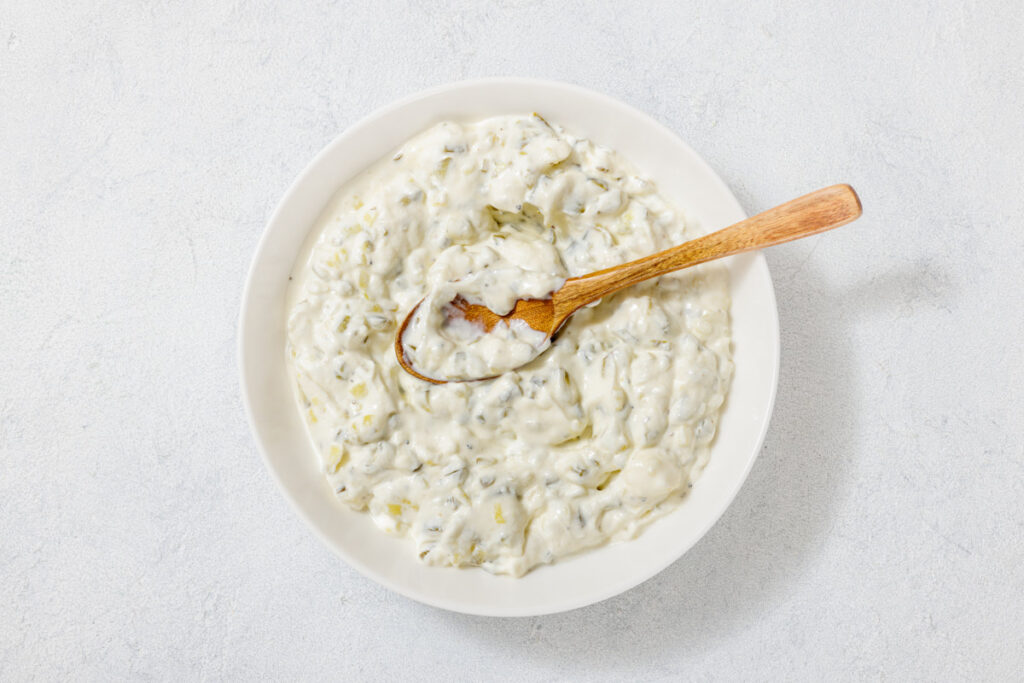 A white bowl filled with creamy tzatziki sauce. A wooden spoon is placed in the center, resting on the rim. The surface shows visible cucumber and herb bits.