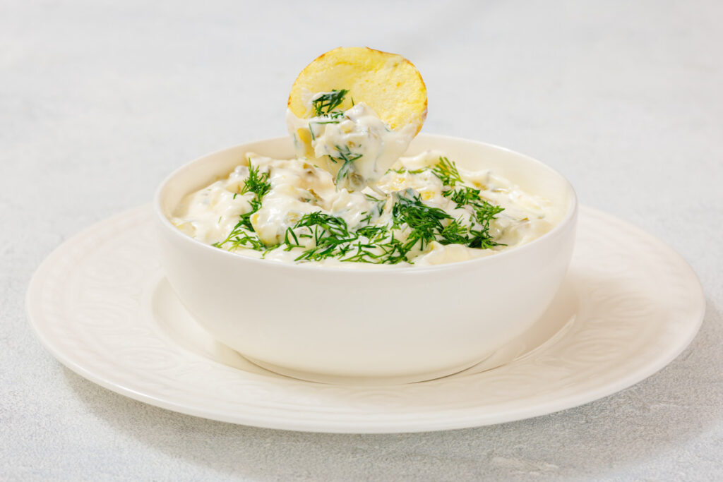 A bowl of Dill Pickle Dip garnished with fresh dill, with a potato chip dipped into it, placed on a white plate.