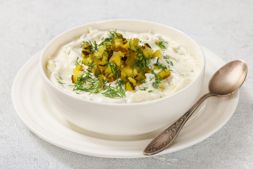 A bowl of Dill Pickle Dip topped with diced pickles and fresh dill, placed on a white plate with a vintage spoon beside it.