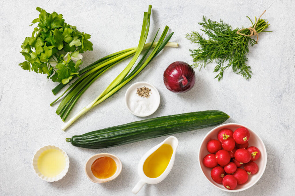 Ingredients on a white surface: radishes, cucumber, red onion, green onions, parsley, dill, lemon juice, honey, olive oil, salt, and pepper.