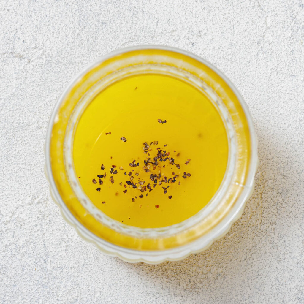 Top view of a clear jar containing yellow liquid with black pepper flakes, set on a textured white surface.