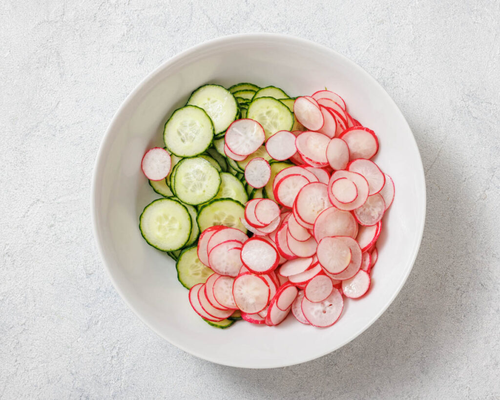 A white bowl contains slices of cucumber on the left and radish on the right, arranged separately on a light textured surface.