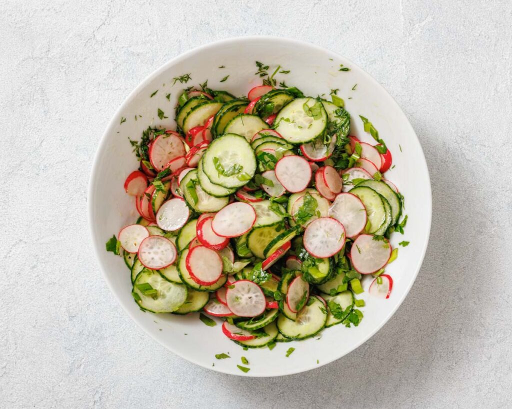 A bowl of sliced cucumbers and radishes mixed with chopped herbs on a light textured surface.