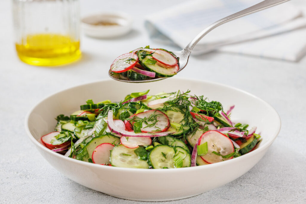 A spoonful of Radish Salad is held above a white bowl filled with the same salad. A jar of oil and a small dish are blurred in the background.