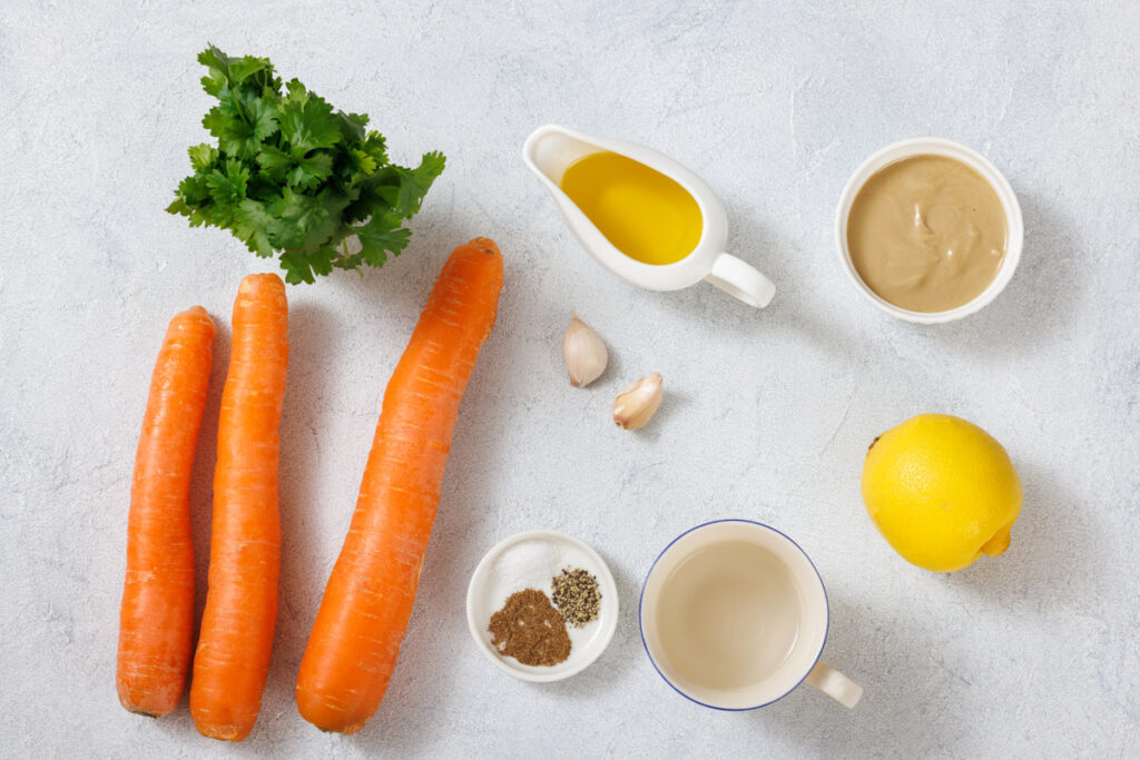 Carrots, cilantro, olive oil, tahini, garlic cloves, lemon, and small bowls of spices and water arranged on a light surface.