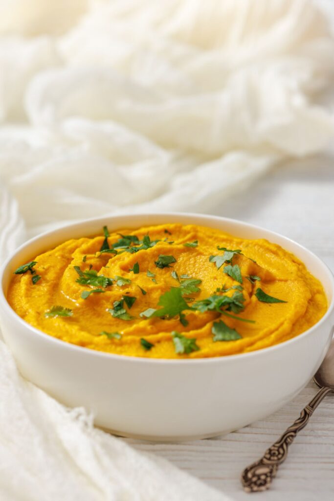 A white bowl filled with Roasted Carrot Hummus, garnished with chopped parsley, on a white surface with a spoon nearby.