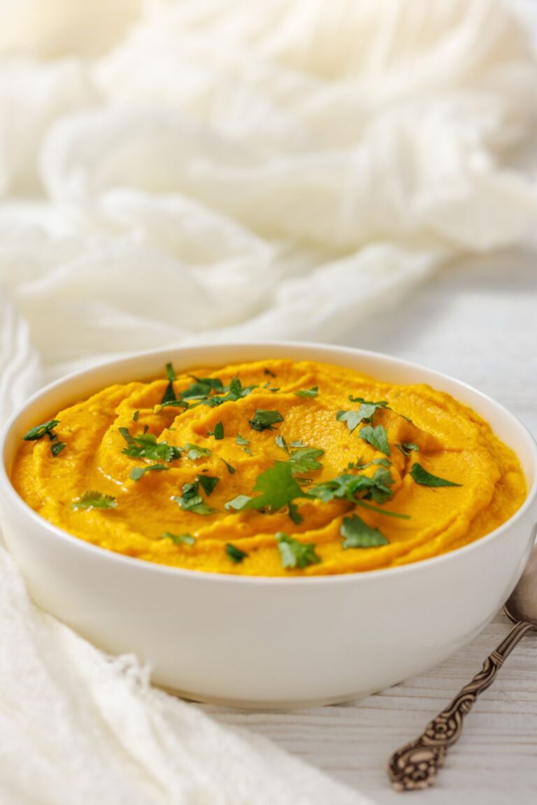 A white bowl filled with Roasted Carrot Hummus, garnished with chopped parsley, on a white surface with a spoon nearby.