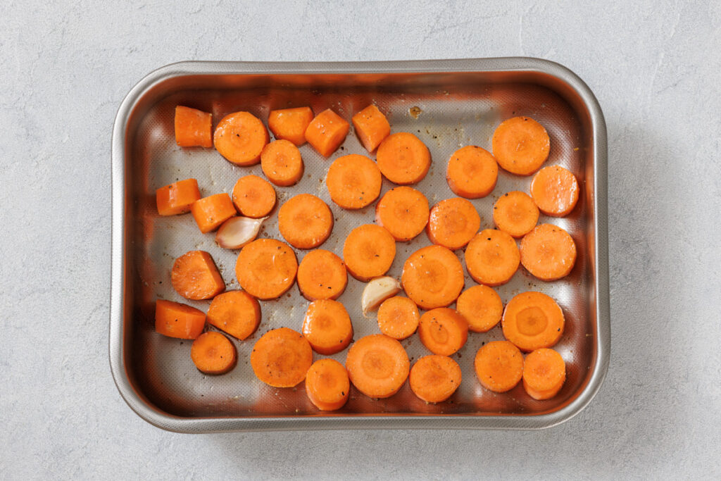 Sliced carrots and garlic pieces seasoned and placed in a metal baking tray on a gray surface.