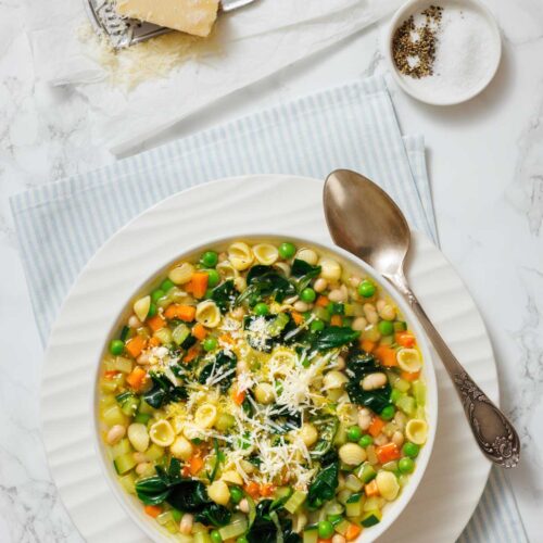 A bowl of Spring Minestrone with pasta, spinach, peas, carrots, and white beans, garnished with grated cheese. A spoon, grated cheese, grater, and pepper are nearby on a striped cloth.