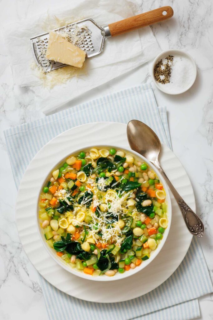 A bowl of Spring Minestrone with pasta, spinach, peas, carrots, and white beans, garnished with grated cheese. A spoon, grated cheese, grater, and pepper are nearby on a striped cloth.