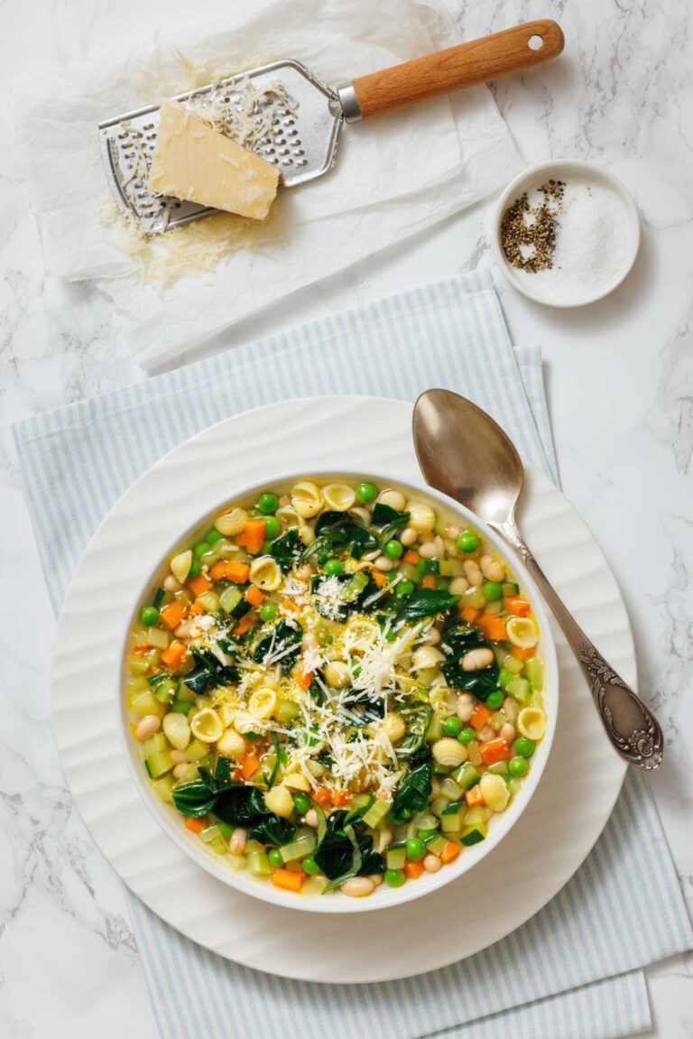 A bowl of Spring Minestrone with pasta, spinach, peas, carrots, and white beans, garnished with grated cheese. A spoon, grated cheese, grater, and pepper are nearby on a striped cloth.