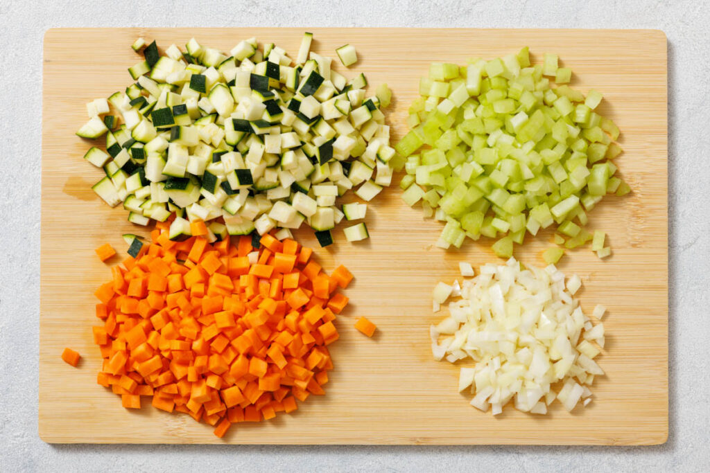 Chopped vegetables on a wooden board include diced zucchini, celery, carrots, and onions arranged separately in four sections.
