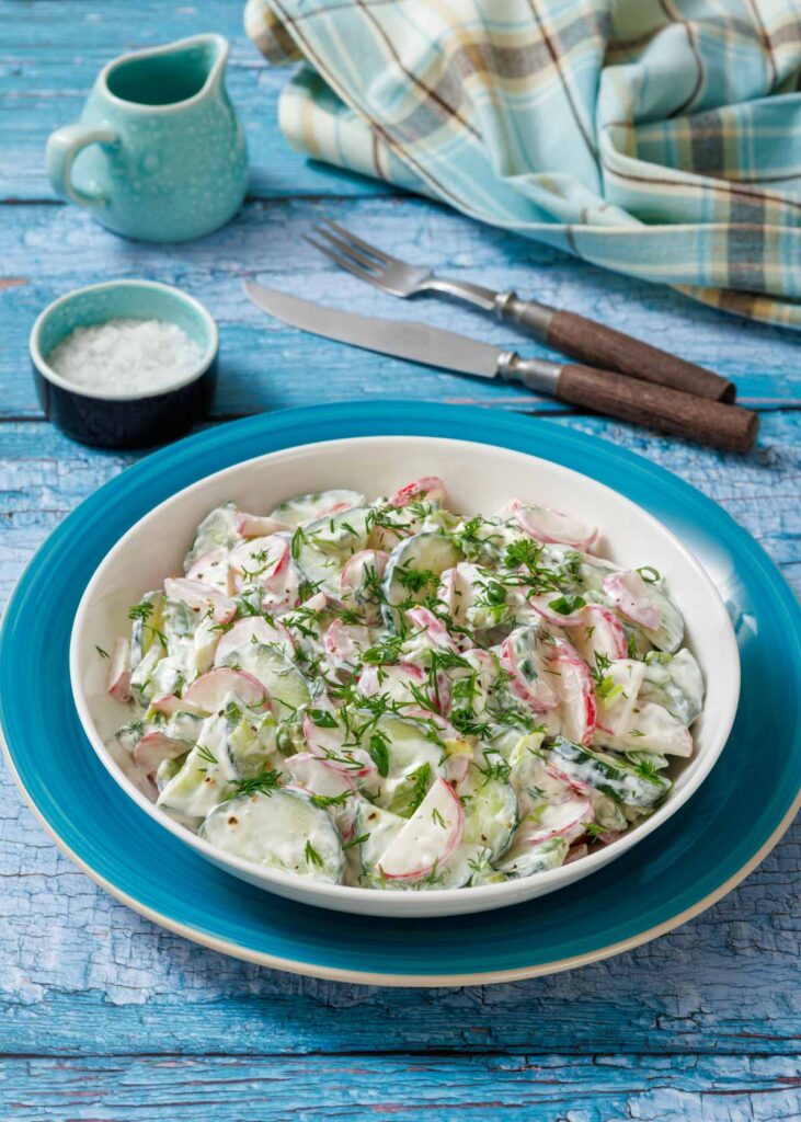 A bowl of creamy cucumber and radish salad with dill on a blue plate. Cutlery and a napkin are in the background on a rustic blue wooden table.