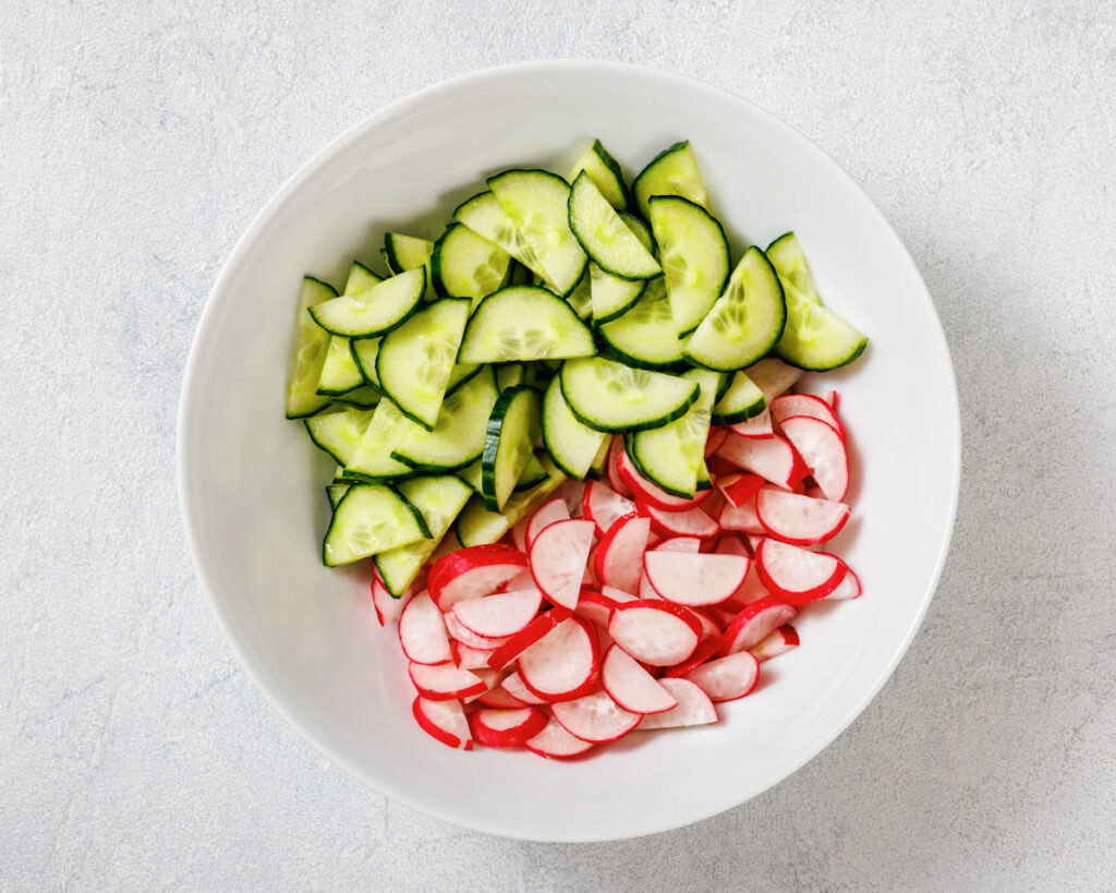 A white bowl containing sliced cucumbers and radishes placed on a light gray surface.