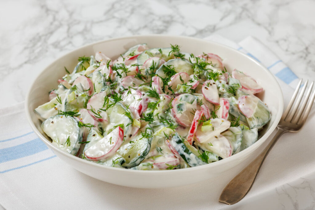 A bowl of creamy cucumber and radish salad garnished with dill, placed on a white and blue cloth. A fork lies beside the bowl.