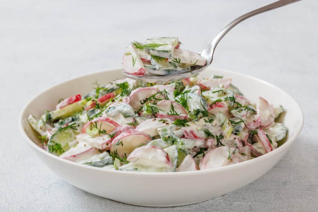 A bowl of Creamy Radish Salad garnished with dill, with a spoon lifting a portion above the bowl.