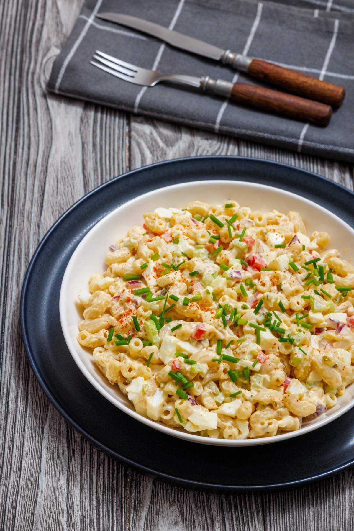 A bowl of macaroni salad with diced vegetables and herbs, garnished with chives. A fork and knife are placed on a checkered napkin beside the dish.