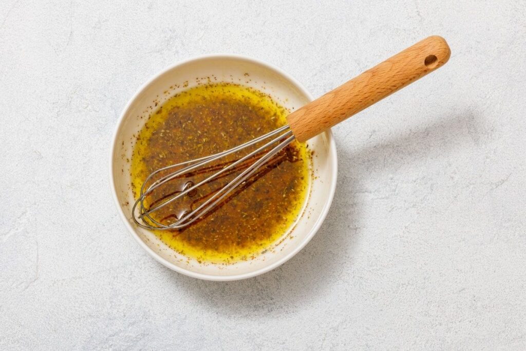 A whisk rests in a bowl containing a mixture of oil and spices on a light-colored surface.