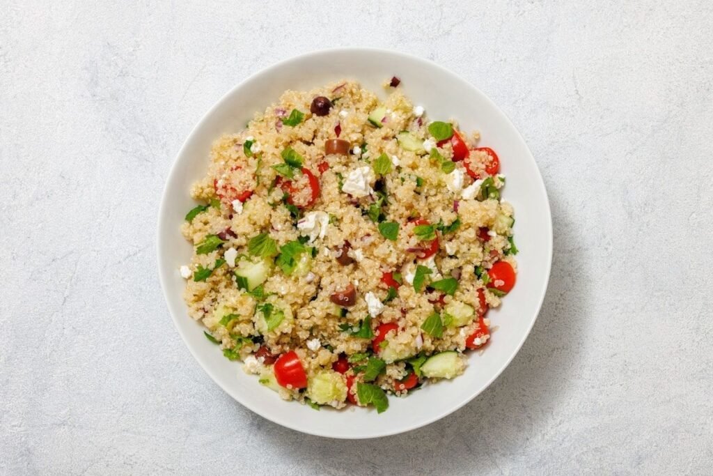 A bowl of quinoa salad with cherry tomatoes, cucumber, feta cheese, olives, and herbs on a light background.