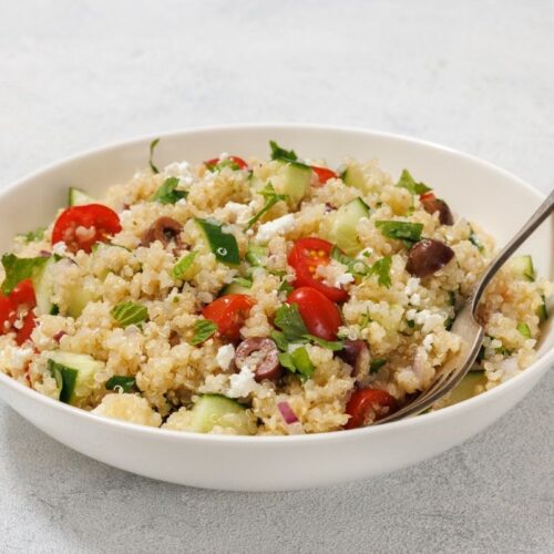 A bowl of quinoa salad with tomatoes, cucumbers, olives, herbs, and feta cheese. A spoon rests in the bowl.