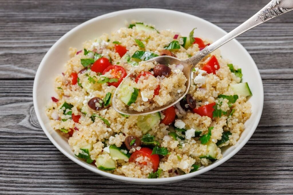 A bowl of quinoa salad with cherry tomatoes, cucumbers, olives, feta cheese, and herbs on a wooden table. A spoon is scooping some salad.