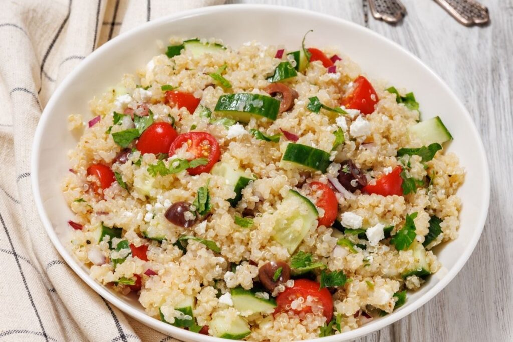 A bowl of quinoa salad with cherry tomatoes, cucumbers, olives, red onions, and feta cheese, garnished with herbs.