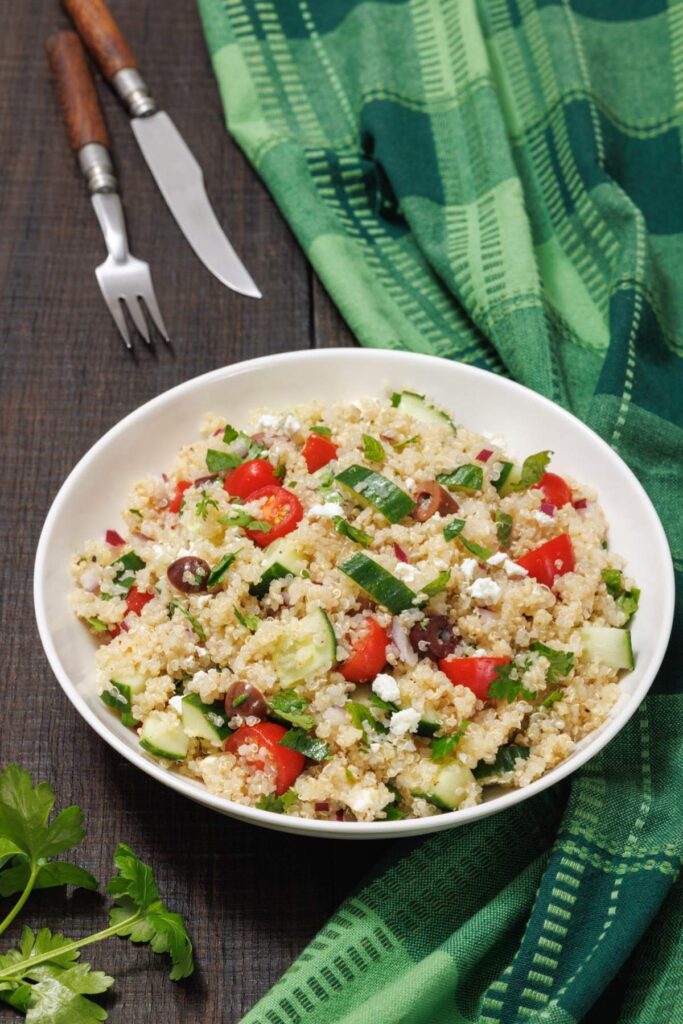 A bowl of quinoa salad with tomatoes, cucumbers, olives, and feta cheese. A fork and knife are beside the bowl on a wooden table with a green plaid cloth.