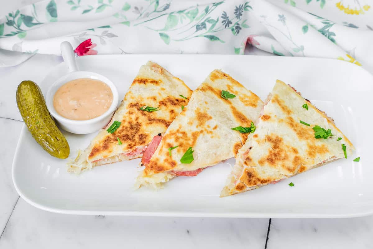 Three Reuben quesadilla slices with herbs on a white plate, accompanied by a pickle and a small dish of dipping sauce.