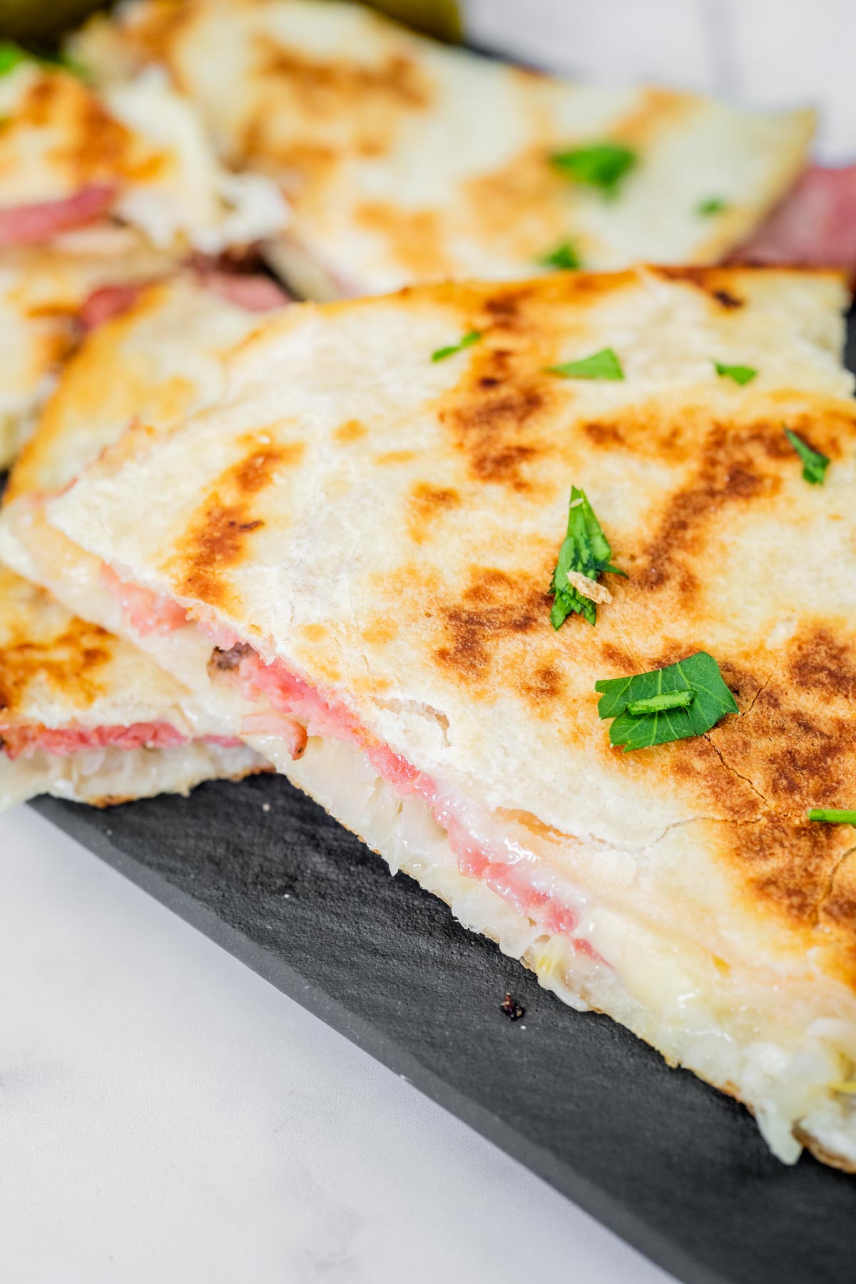Close-up of a quesadilla with melted cheese and ham on a slate board, garnished with chopped parsley.