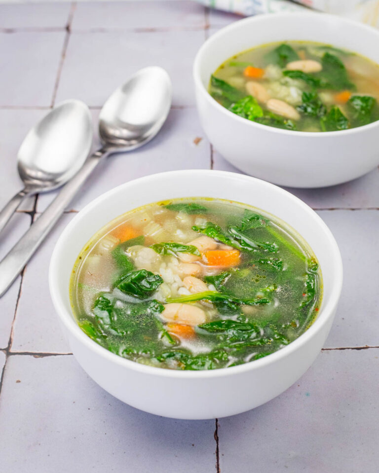 A close-up of a white bowl filled with rice and white bean soup, featuring vibrant spinach leaves, diced carrots, and beans in a clear broth. Another bowl of soup and two silver spoons rest on a tiled surface in the background.