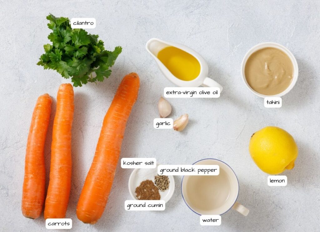 Ingredients for Roasted Carrot Hummus recipe arranged on a surface: cilantro, carrots, extra-virgin olive oil, tahini, garlic, kosher salt, ground cumin, ground black pepper, water, and a lemon.