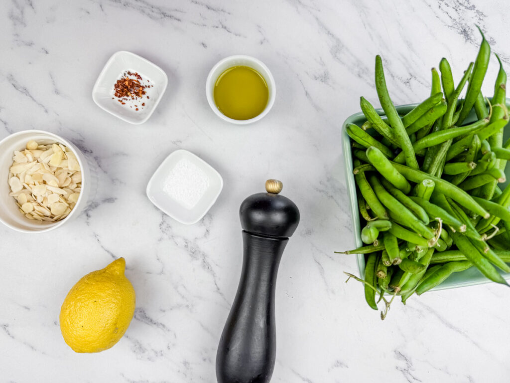 Assorted ingredients on a marble surface: fresh green beans, sliced almonds, lemon, olive oil, red pepper flakes, salt, and a black pepper grinder.